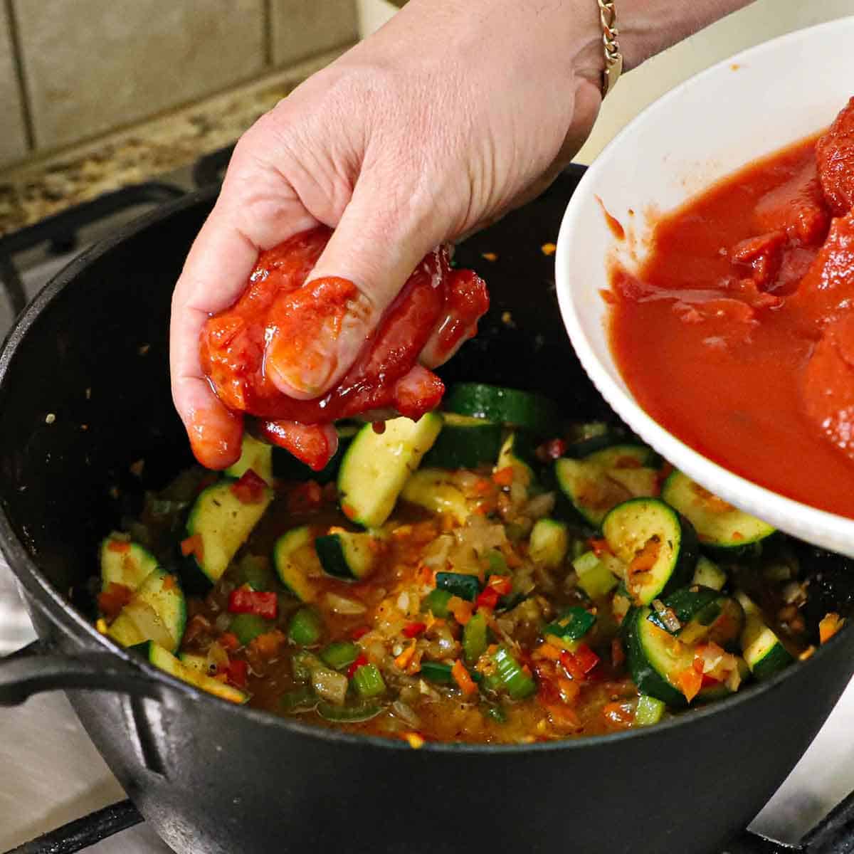 A person squeezing canned whole tomatoes with one hand over a large pot filled with simmering seasoned vegetables while holding a dish of whole tomatoes in his other hand.