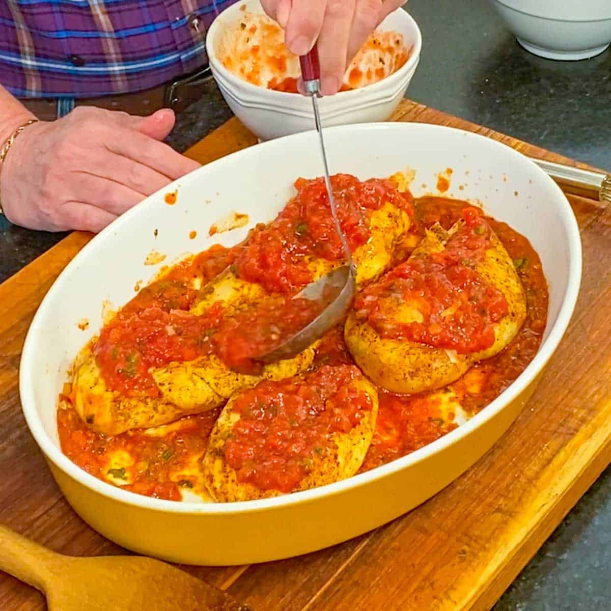 A person using a ladle to pour homemade salsa over the tops of seared seasoned chicken breasts in a large oval casserole dish on a wooden cutting board.