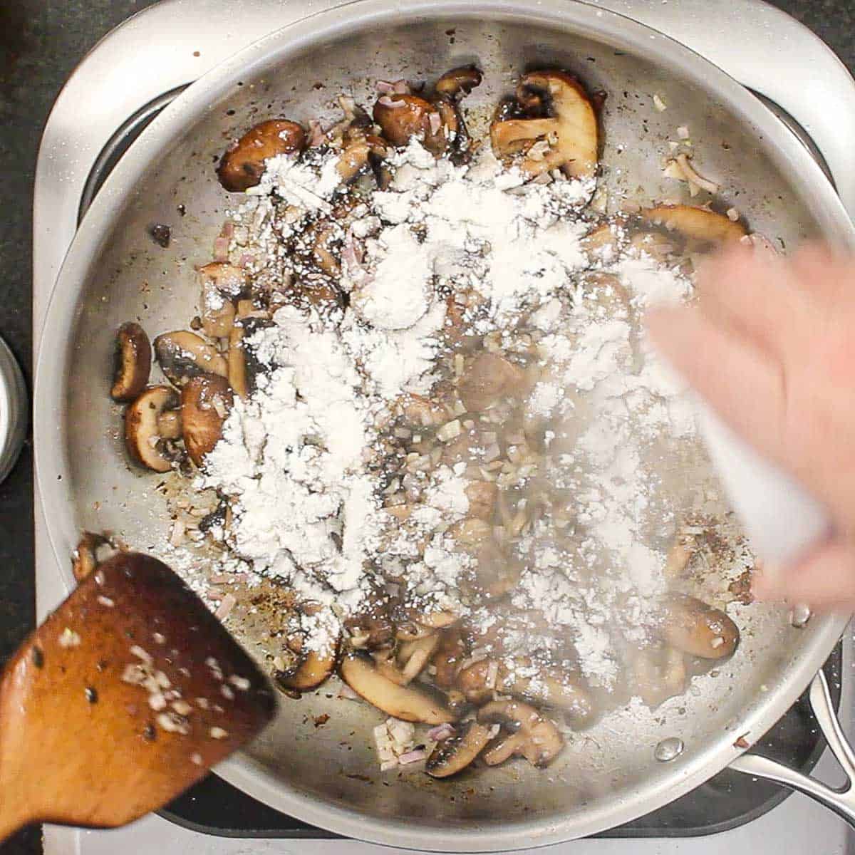 A person pouring all-purpose flour into a skillet that is filled with sautéed sliced mushrooms and shallots.