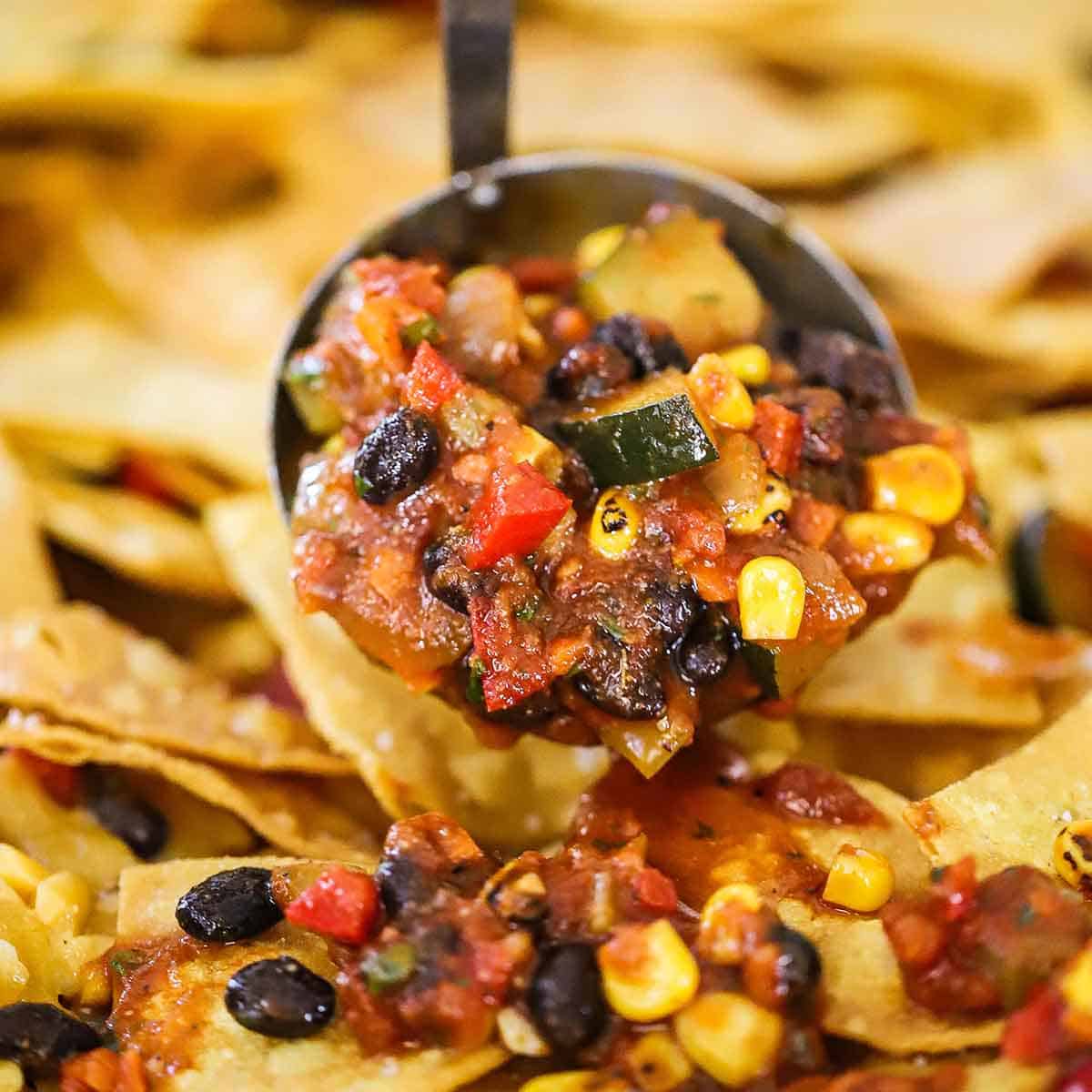 A person using a ladle to place vegetarian and black bean chili over the tops of fried tortilla chips on a large sheet pan.