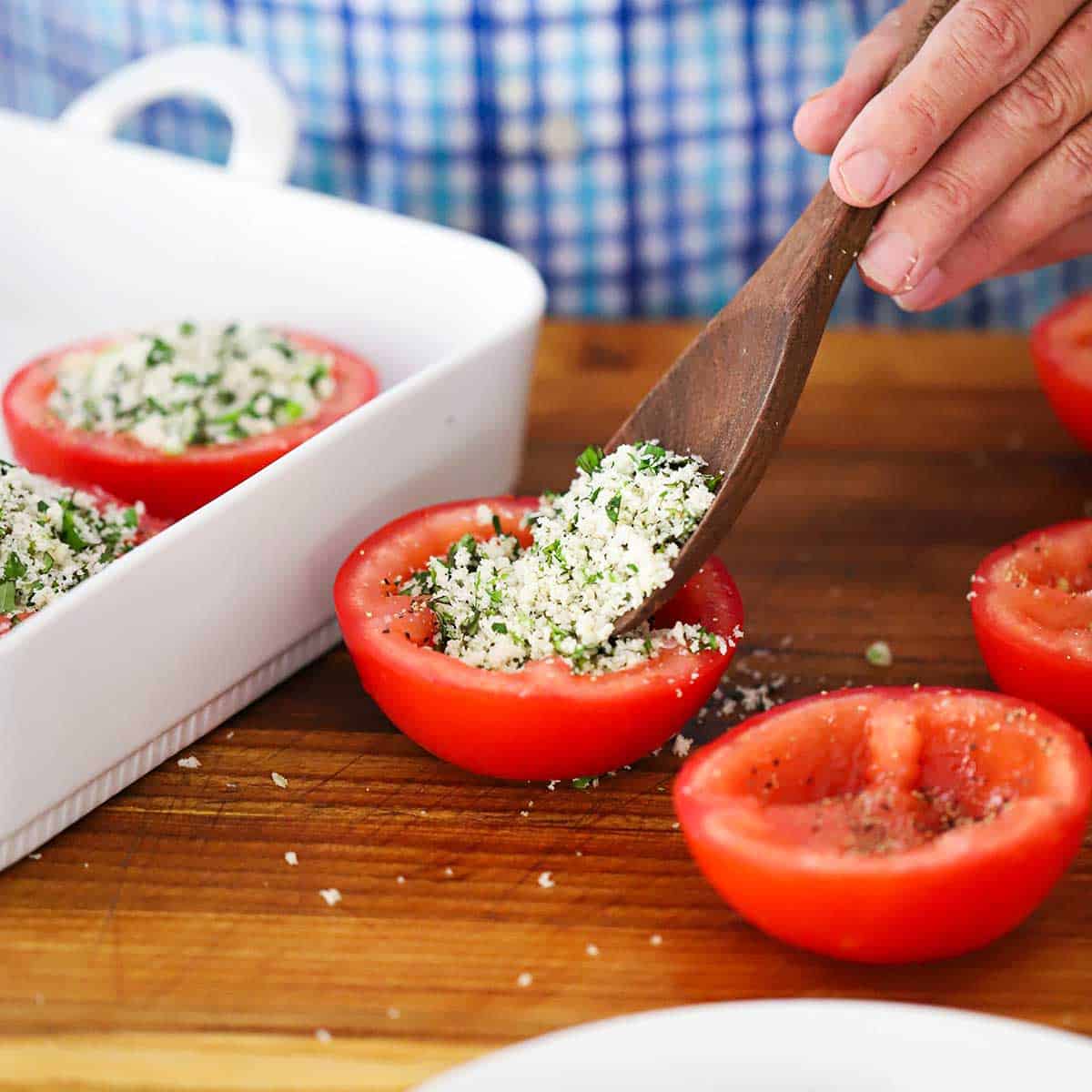A person using a wooden spoon to fill the a hollowed out red tomato half with an herb breadcrumb mixture.