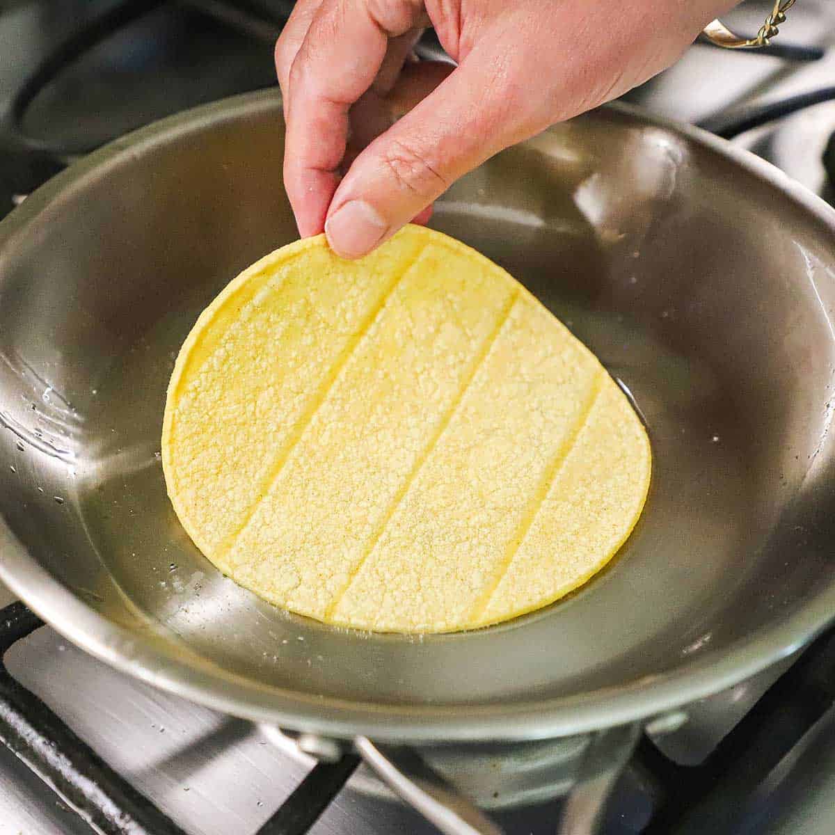 A person placing a corn tortilla into a small steel skillet that has a thin layer of hot oil in the bottom of it on a gas stove.