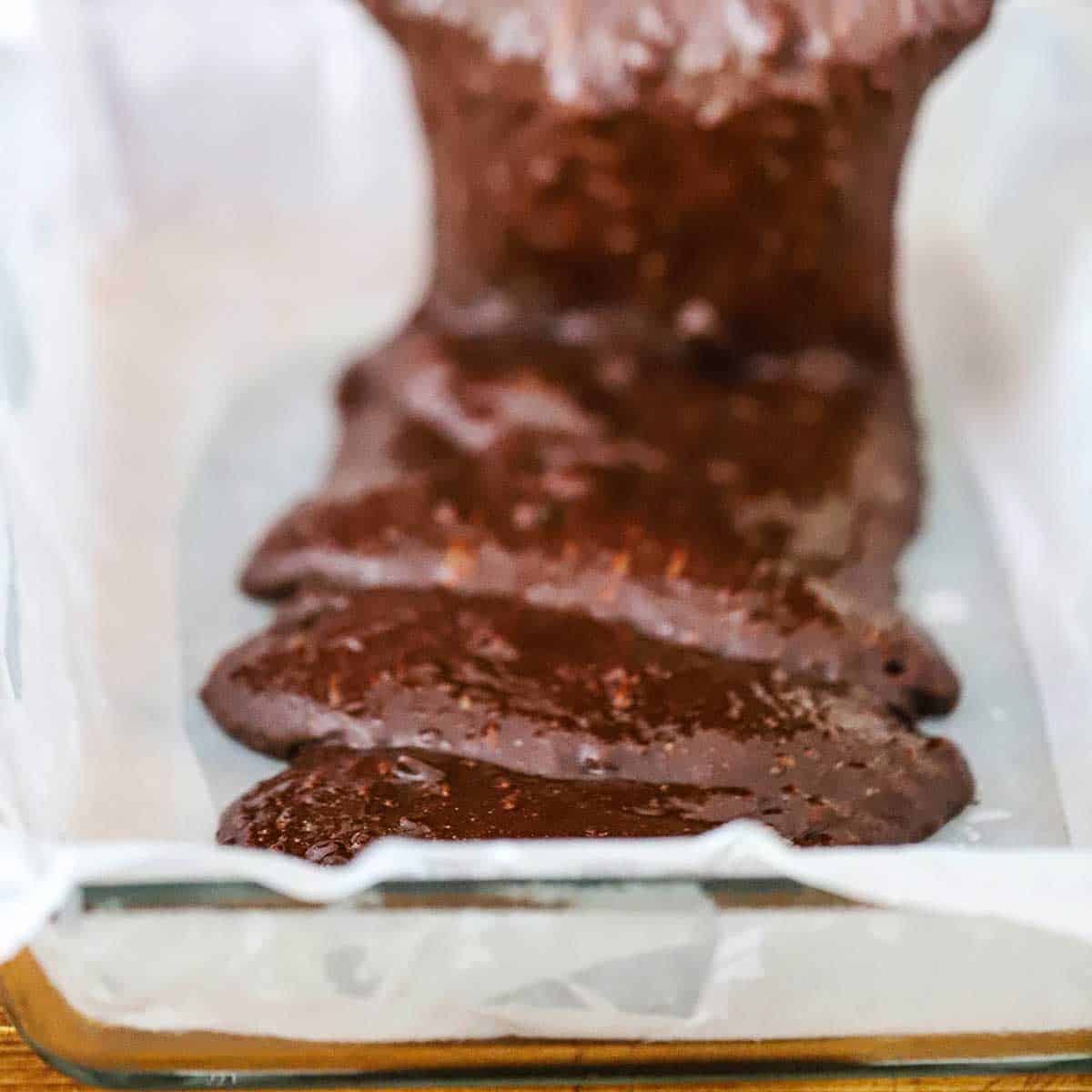 A person pouring fudge brownie batter into a baking dish that is lined with parchment paper.