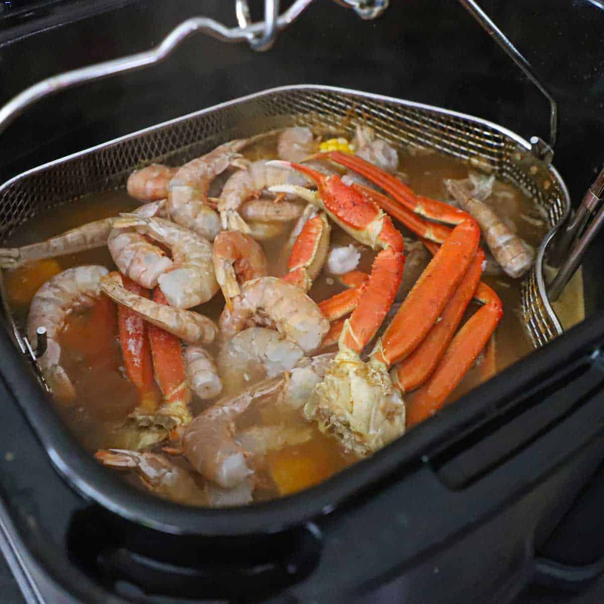 A person lowering a large basket filled with shrimp and snow crab legs into a pot filled with a simmering seafood boil.