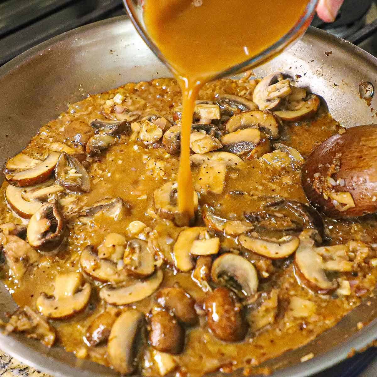 A person pouring a mixture of beef stock and Dijon mustard from a glass measuring cup into a large skillet filled with the simmering liquid and sautéed mushrooms and shallots.