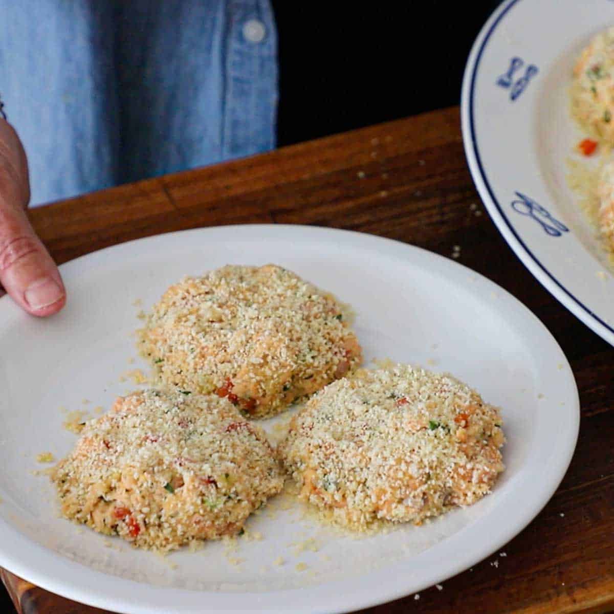 A person holding a plate filled with three uncooked fresh salmon cakes on a cutting board.