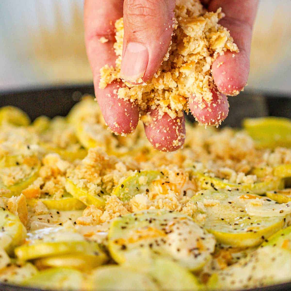 A person using his hand to sprinkle a crushed cracker and butter mixture over the top of an uncooked summer squash casserole in a large cast-iron skillet.