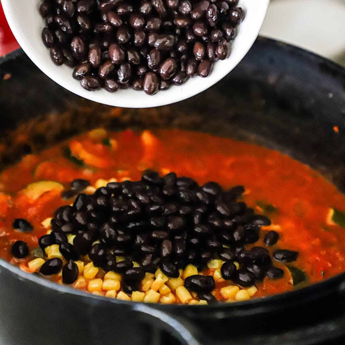 A person transferring tender black beans from a white bowl into a large black pot filled with a vegetable chili and corn kernels.