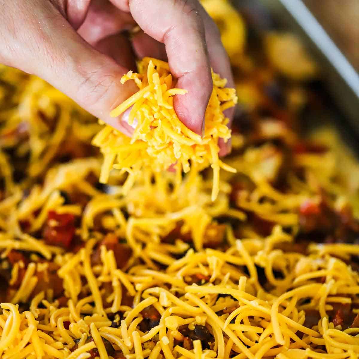 A person using his fingers to sprinkle shredded cheddar cheese over the top of sheet pan nachos.