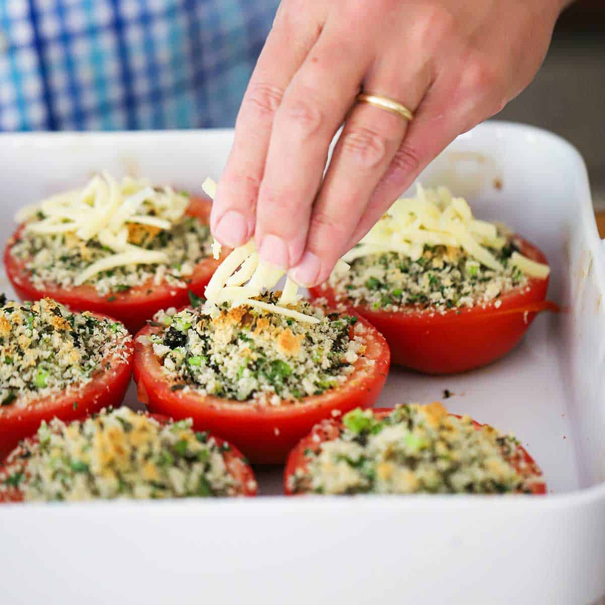 A person placing shredded Gruyere cheese over the tops of baked stuffed red tomatoes that are in a square white baking dish.