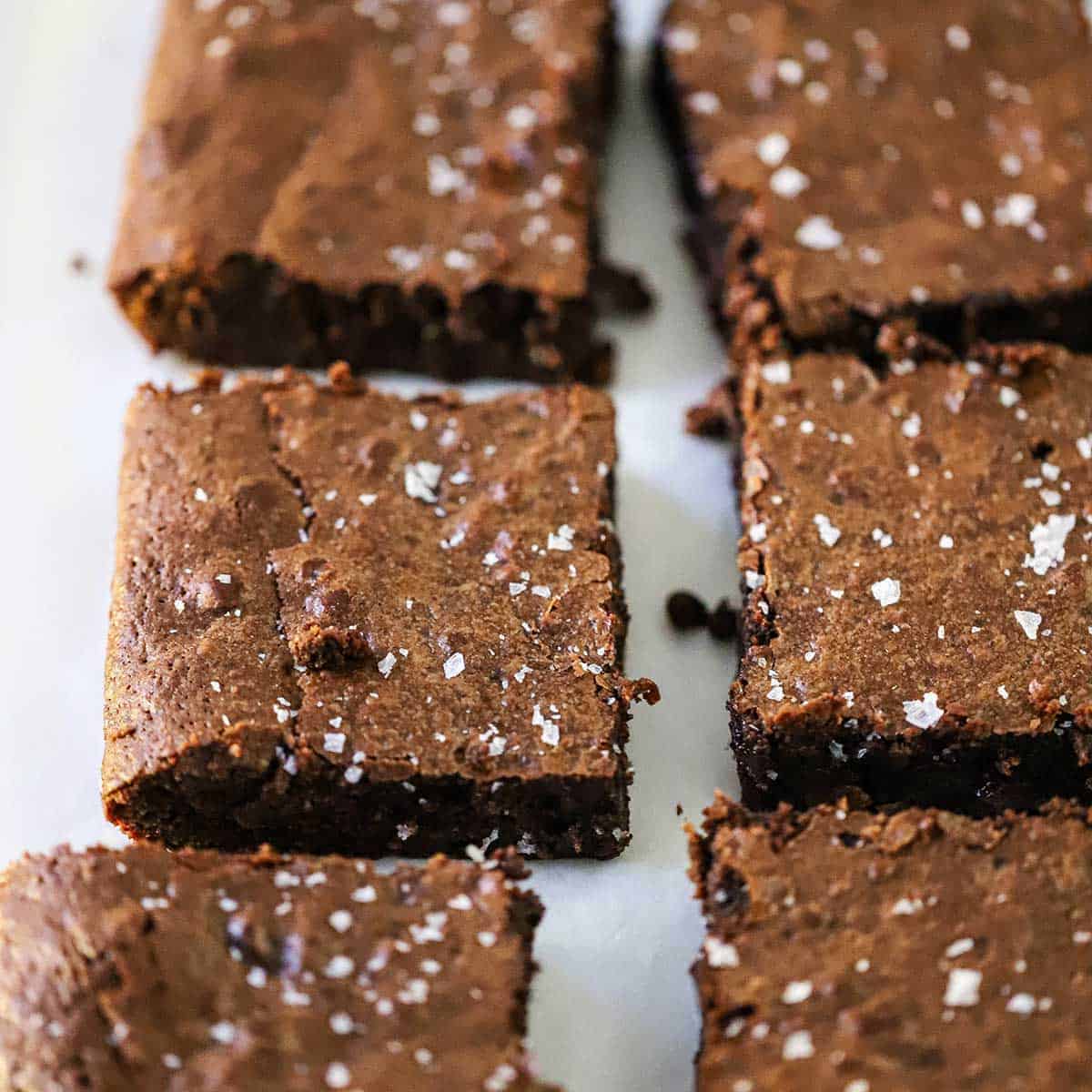 Three rows of square fudge brownies that are topped with coarse sea salt all resting on a sheet of parchment paper.