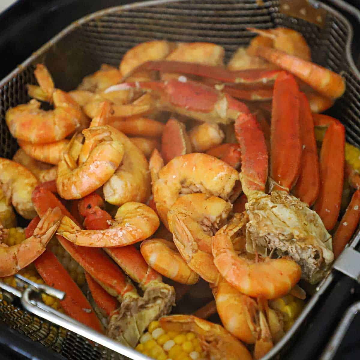 A close-up view of a large basket that has been raised from boiling Cajun liquid and is filled with cooked shrimp, crab legs, corn on the cob, sausage, and baby potatoes.