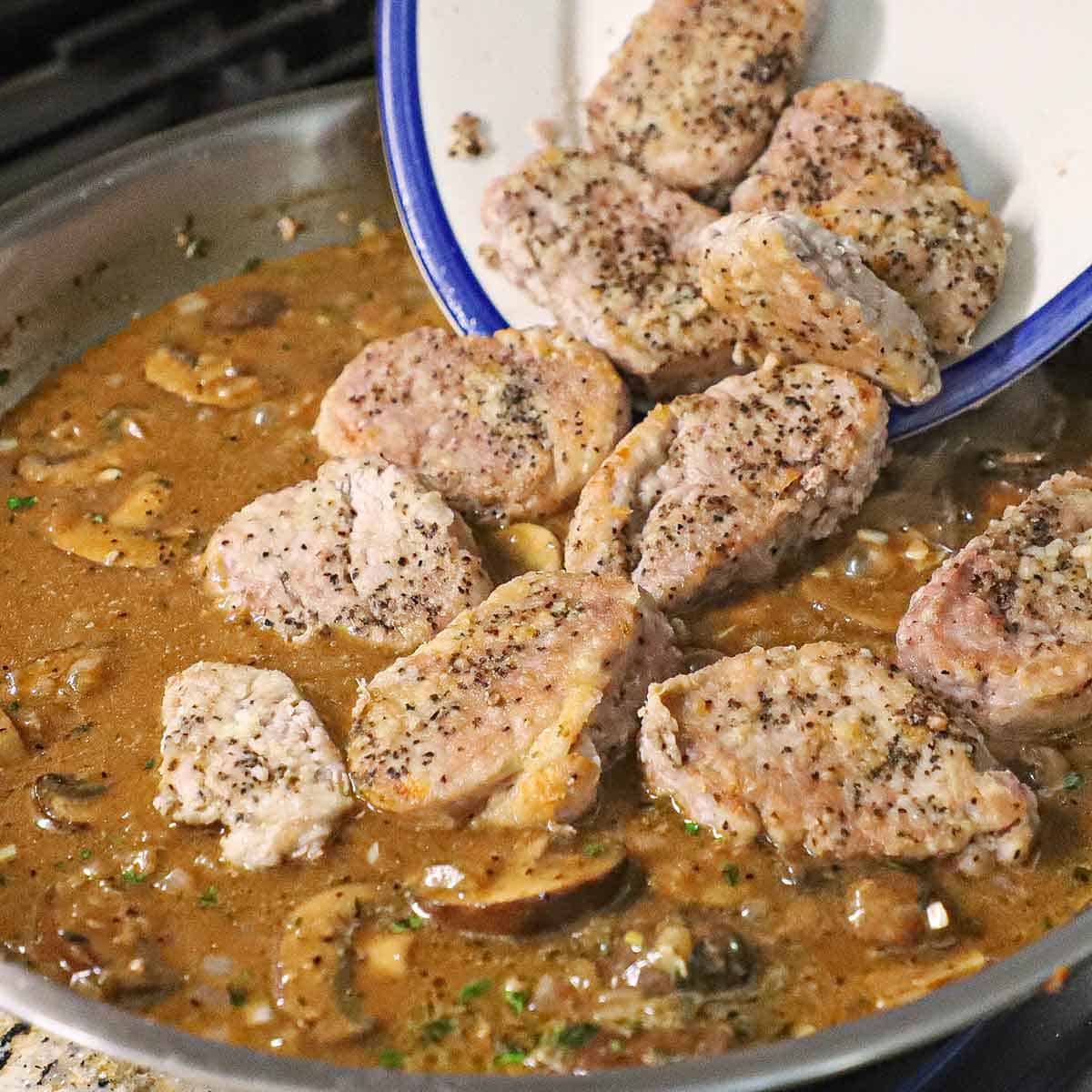 A person transferring seared seasoned pork medallions from a large platter into a skillet filled with a brown Dijon mushroom sauce.