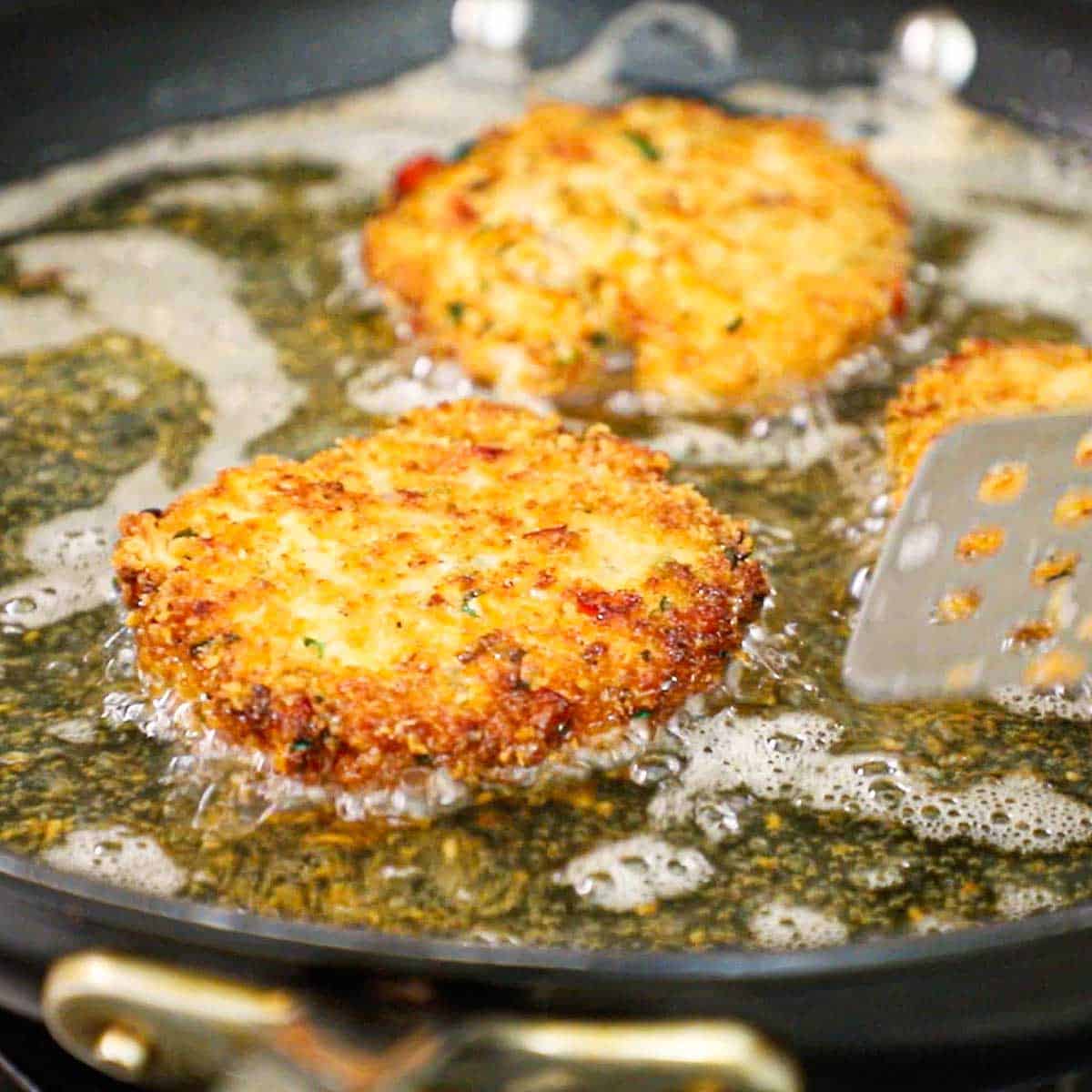 A close-up view of three fresh salmon cakes being lightly fried in vegetable oil in a black large skillet on a gas stove.