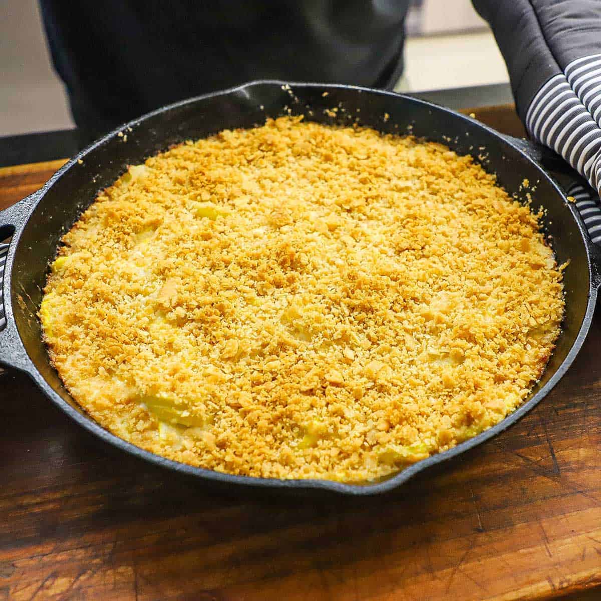 A person wearing oven mitts holding the handles of a large cast-iron skillet filled with a freshly baked summer squash casserole.