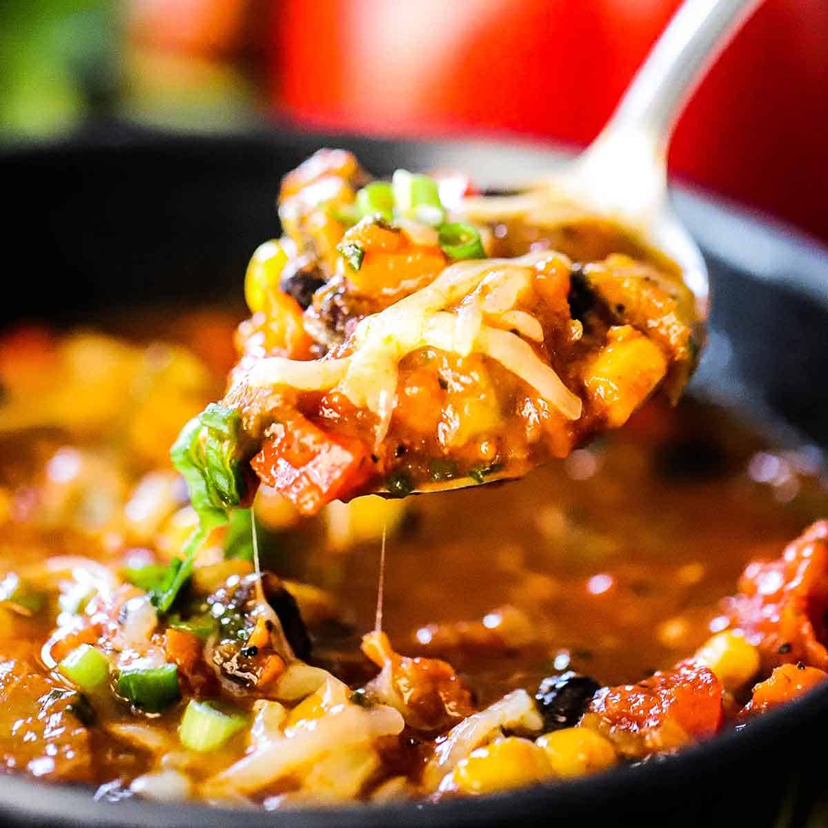 A spoon being used to raise a large spoonful of Vegetable Chili with Black Beans topped with melty cheese from a pot of the same.