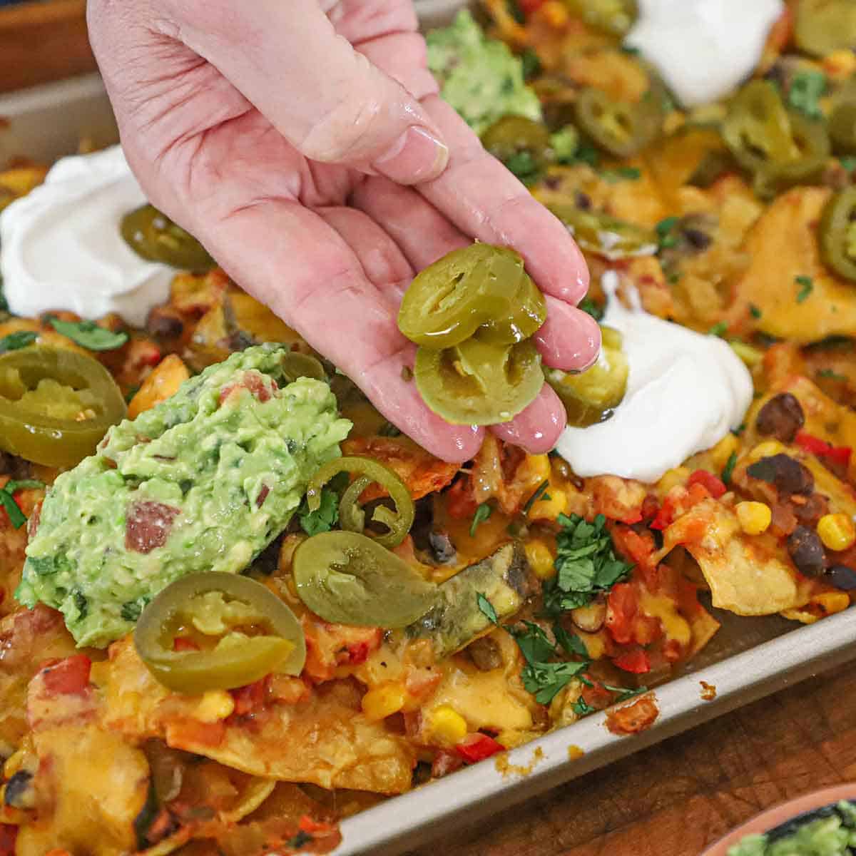 A person holding sliced pickled jalapeños over a large serving of sheet pan nachos with dollops of sour cream and guacamole on the nachos.