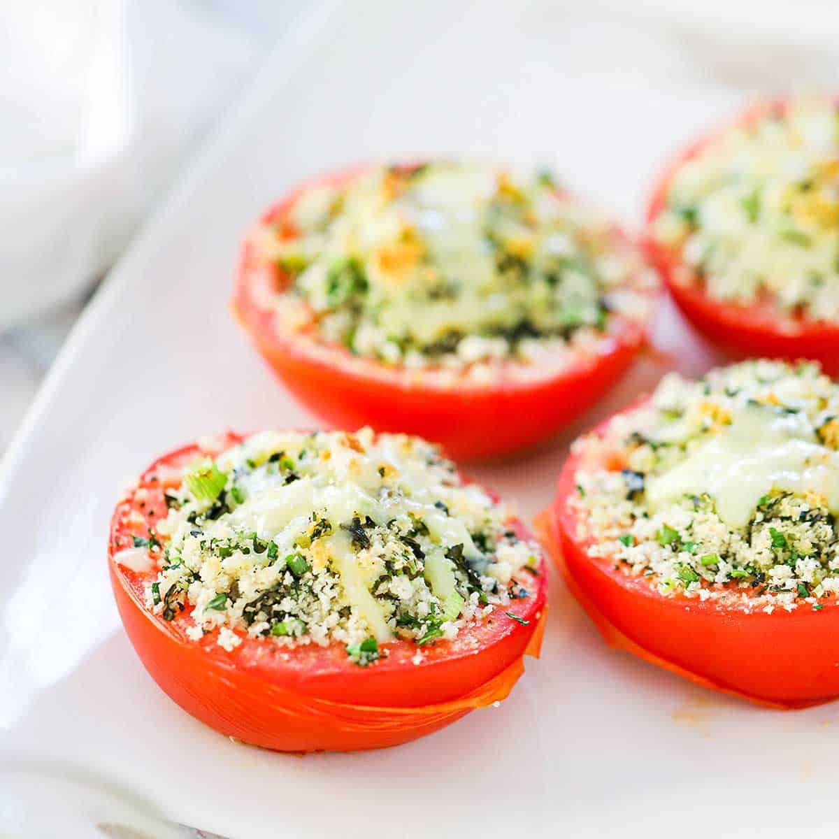 A close-up view of four baked stuffed tomatoes that are lightly browned on top and resting on a white serving platter.