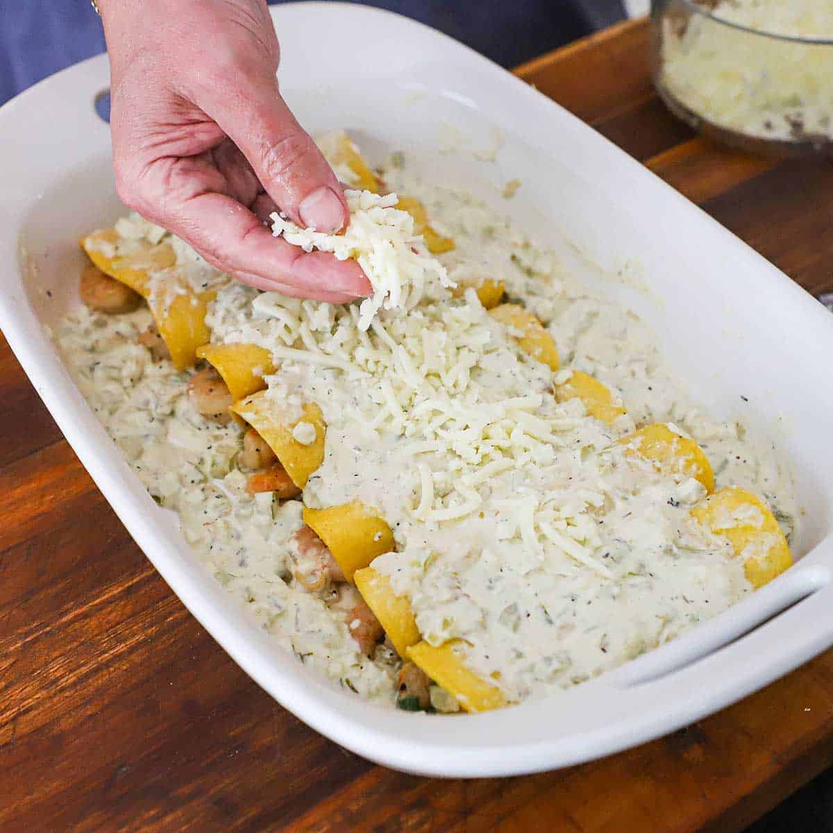 A person sprinkling shredded Monterey Jack cheese over the top of a line of Creamy Shrimp Enchiladas in a baking dish.