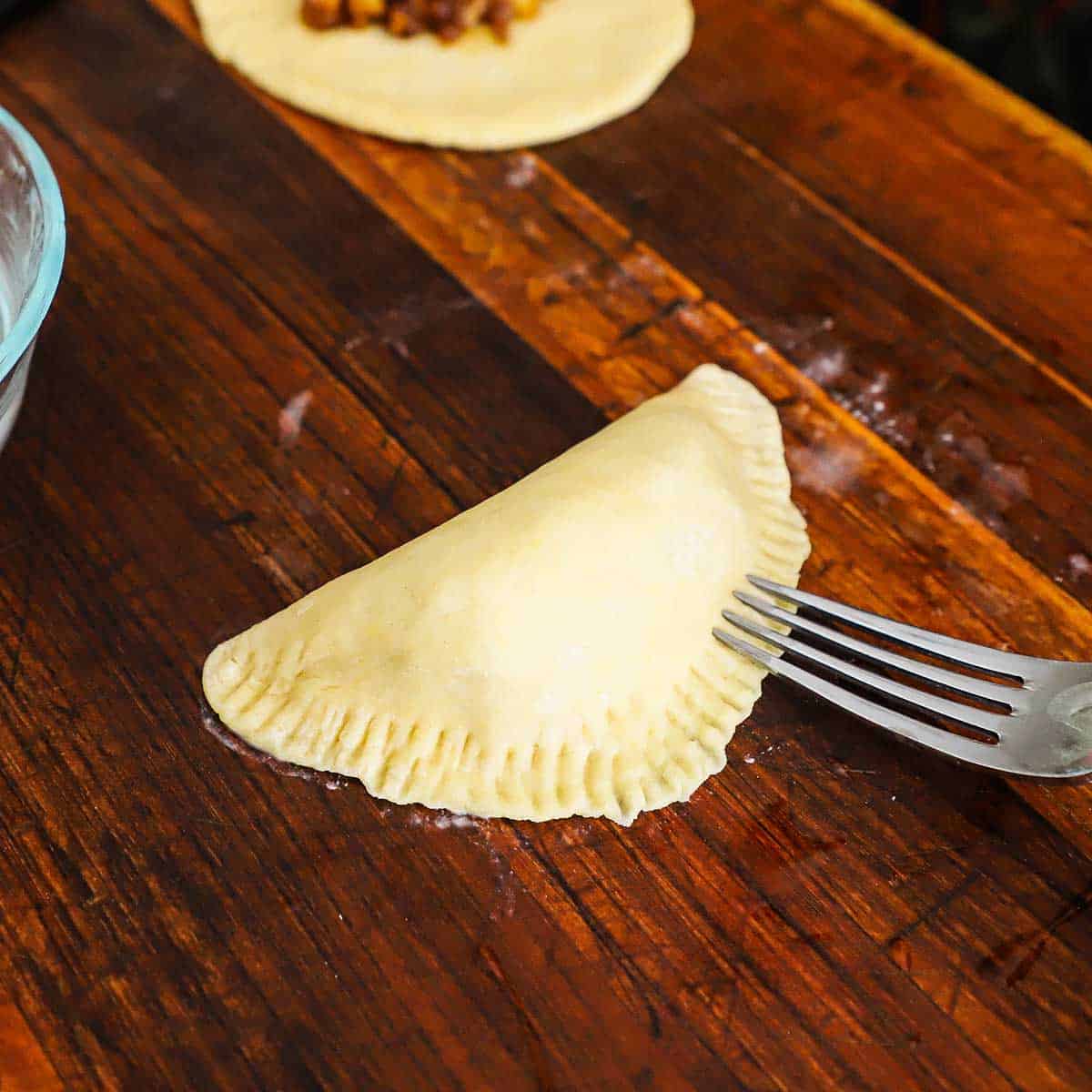 A person using the ends of a fork to crimp the edges of an uncooked fruit pie on a wooden cutting board.