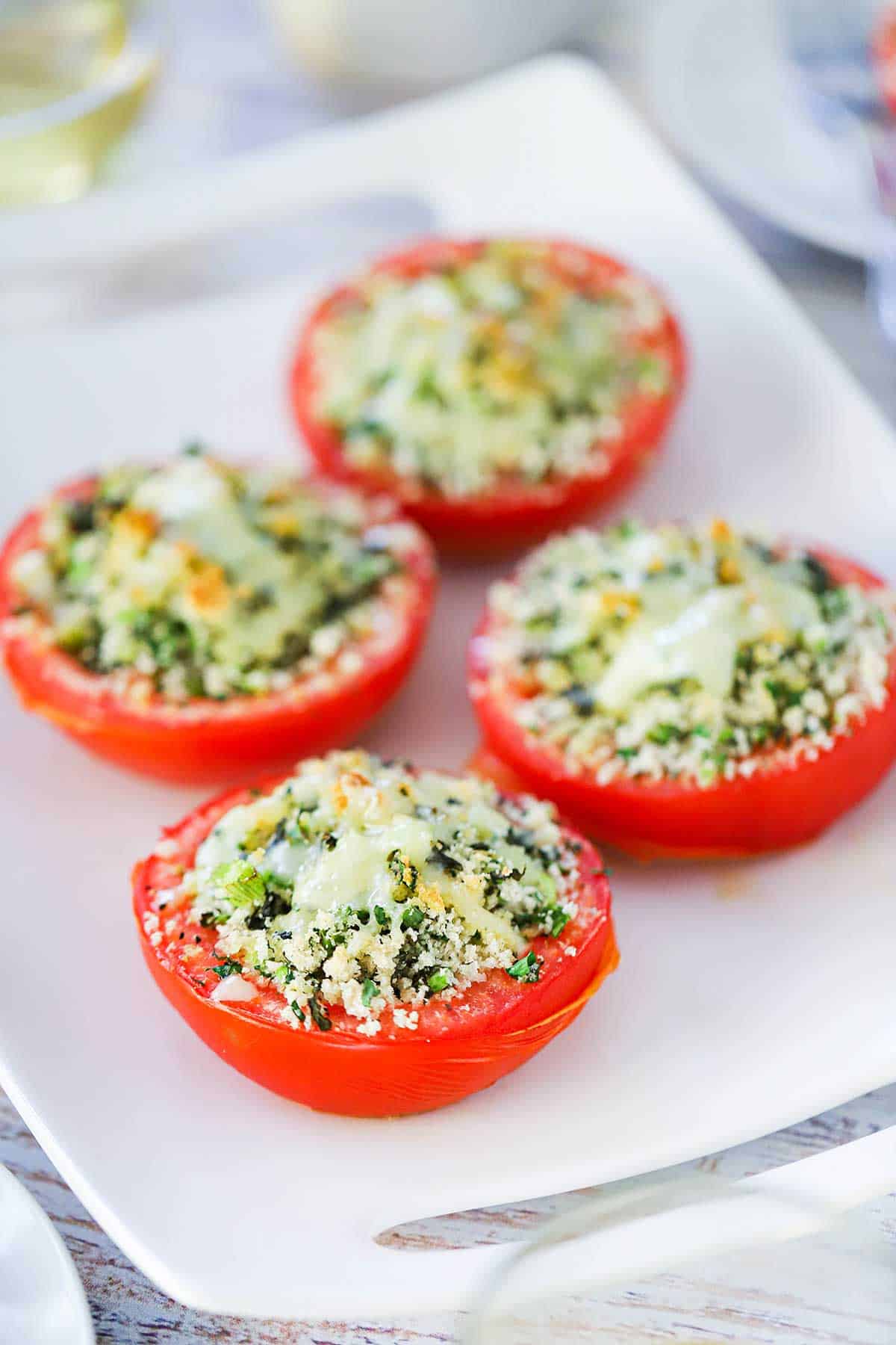 A straight-on view of four baked stuffed summer tomatoes that are topped with lightly browned melted cheese all on a white serving platter.