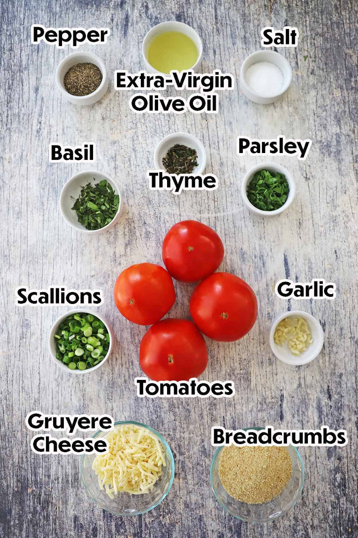 An arrangement of ingredients for Baked Stuffed Summer Tomatoes on a grey wooden background including red tomatoes, garlic, breadcrumbs, Gruyere cheese, scallions, fresh herbs, and seasonings.