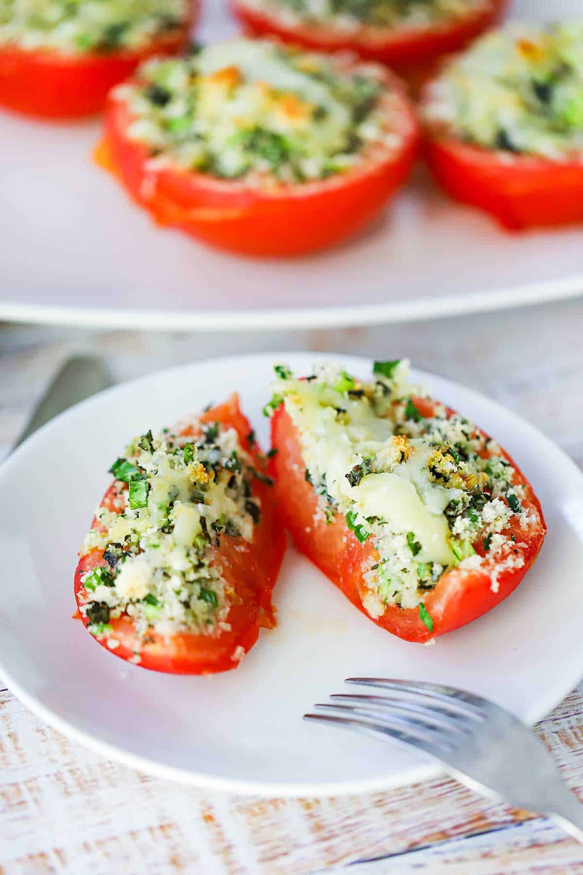 A straight-on view of a Baked Stuffed Summer Tomato that has been cut in half on a small white plate with a fork resting on the edge of the plate.