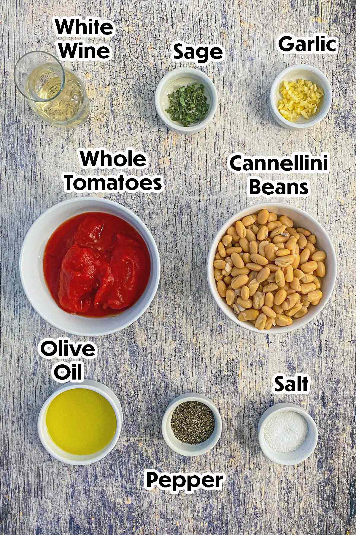 An arrangement of ingredients for Sautéed White Beans with Garlic, Sage, and Tomatoes on a grey wooden background including cannellini beans, whole tomatoes, garlic, wine, oil, sage, and seasonings.
