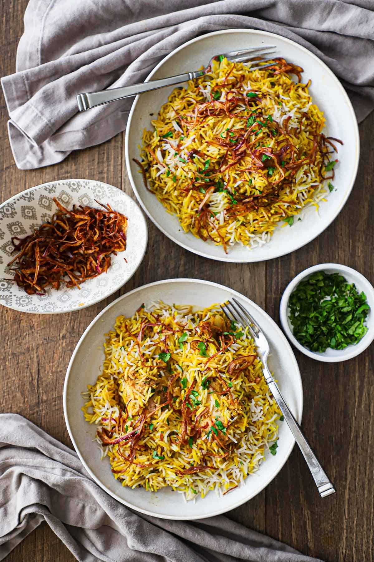 An overhead view of two side-by-side shallow bowls both filled with servings of Hyderabadi Chicken Biryani with a small bowl of fried onions on one side and another small bowl filled with chopped cilantro on the other side.