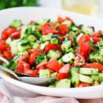 A close-up view of a cucumber tomato salad in a large oval white platter garnished with chopped herbs.