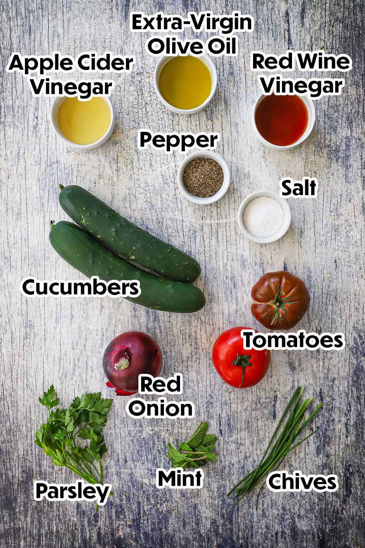 An arrangement of ingredients for Cucumber Tomato Salad on a grey wooden background including cucumbers, tomatoes, red onion, apple cider vinegar, red wine vinegar, oil, seasonings, and fresh herbs.
