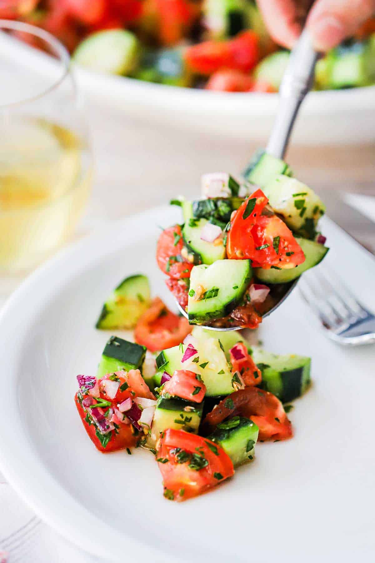 A person using a spoon to place a serving of Chopped Cucumber Tomato Salad onto a white dinner plate next to a glass of white wine.