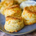 A close-up view of flaky cheddar biscuits sitting in a row on an oval grey platter with a small bowl of softened butter next to the biscuits.