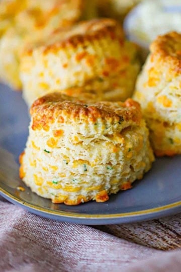 A close-up view of flaky cheddar biscuits sitting in a row on an oval grey platter with a small bowl of softened butter next to the biscuits.
