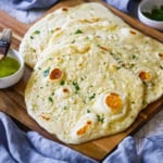 An overhead view of a stack of homemade garlic naan with a small bowl of garlic-cilantro butter nearby as well as a couple of blue cloth napkins.