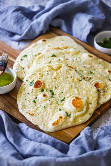 An overhead view of a stack of homemade garlic naan with a small bowl of garlic-cilantro butter nearby as well as a couple of blue cloth napkins.