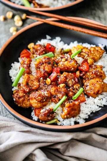 A staight-on view of a black shallow bowl filled with a serving of crispy kung pao cauliflower on top of steamed white rice garnished with strips of scallions and toasted sesame seeds.