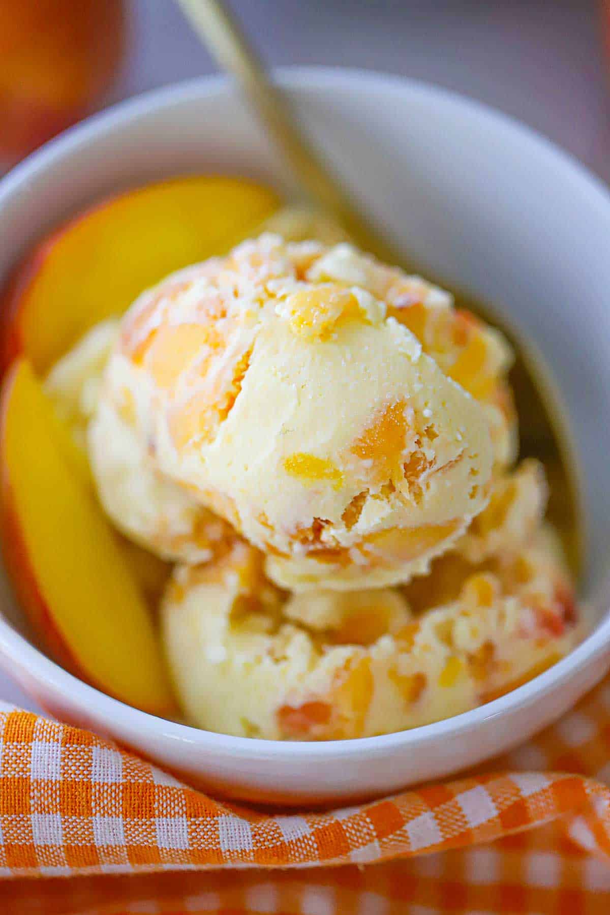 A close-up view of several scoops of homemade peach ice cream with a couple of peach slices all in a white ice cream bowl.