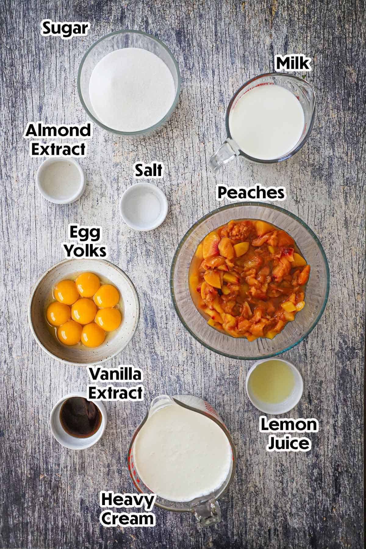 An arrangement of ingredients for homemade peach ice cream on a grey wooden background including chopped fresh peaches, egg yolks, heavy cream, milk, lemon juice, vanilla extract, sugar, salt, and almond extract.
