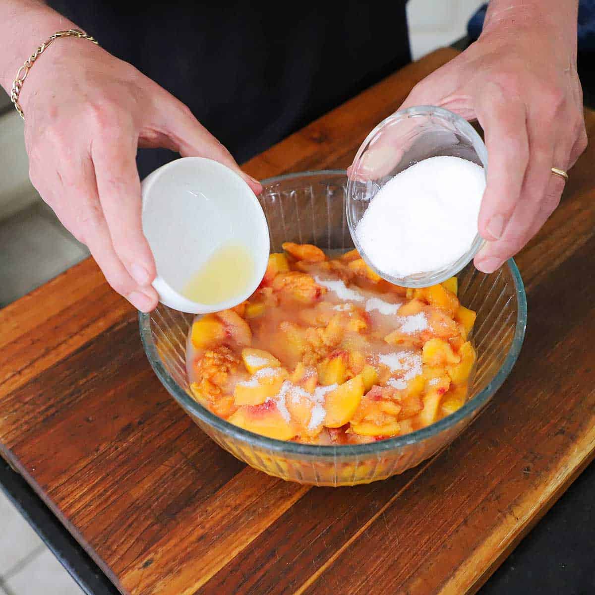 A person holding a small bowl of sugar in one hand and another small bowl filled almond extract in the other hand, both over a larger glass bowl filled with fresh, chopped peaches.