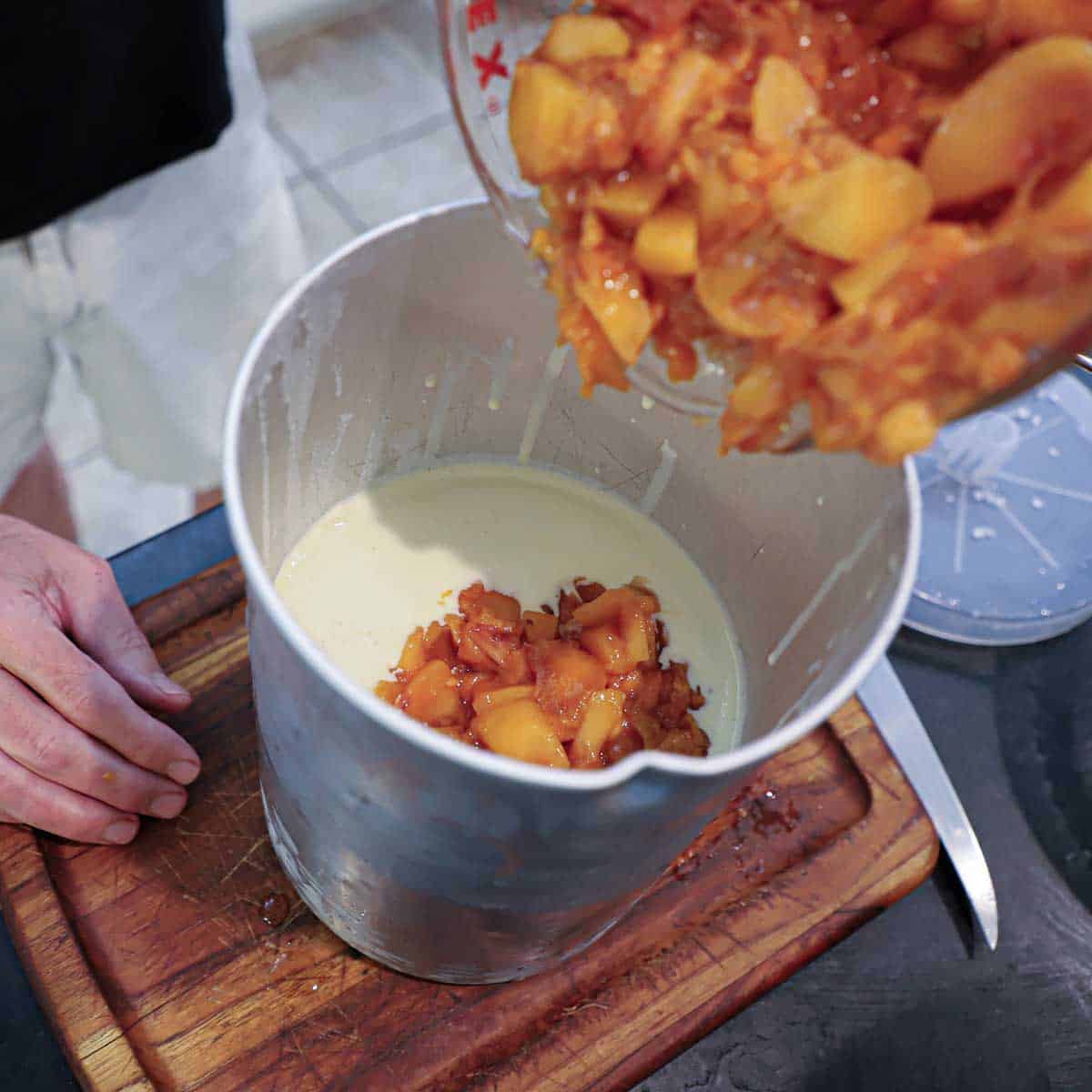 A person transferring chunks of macerated peaces into a canister that is partially filled with freshly churned peach ice cream custard.