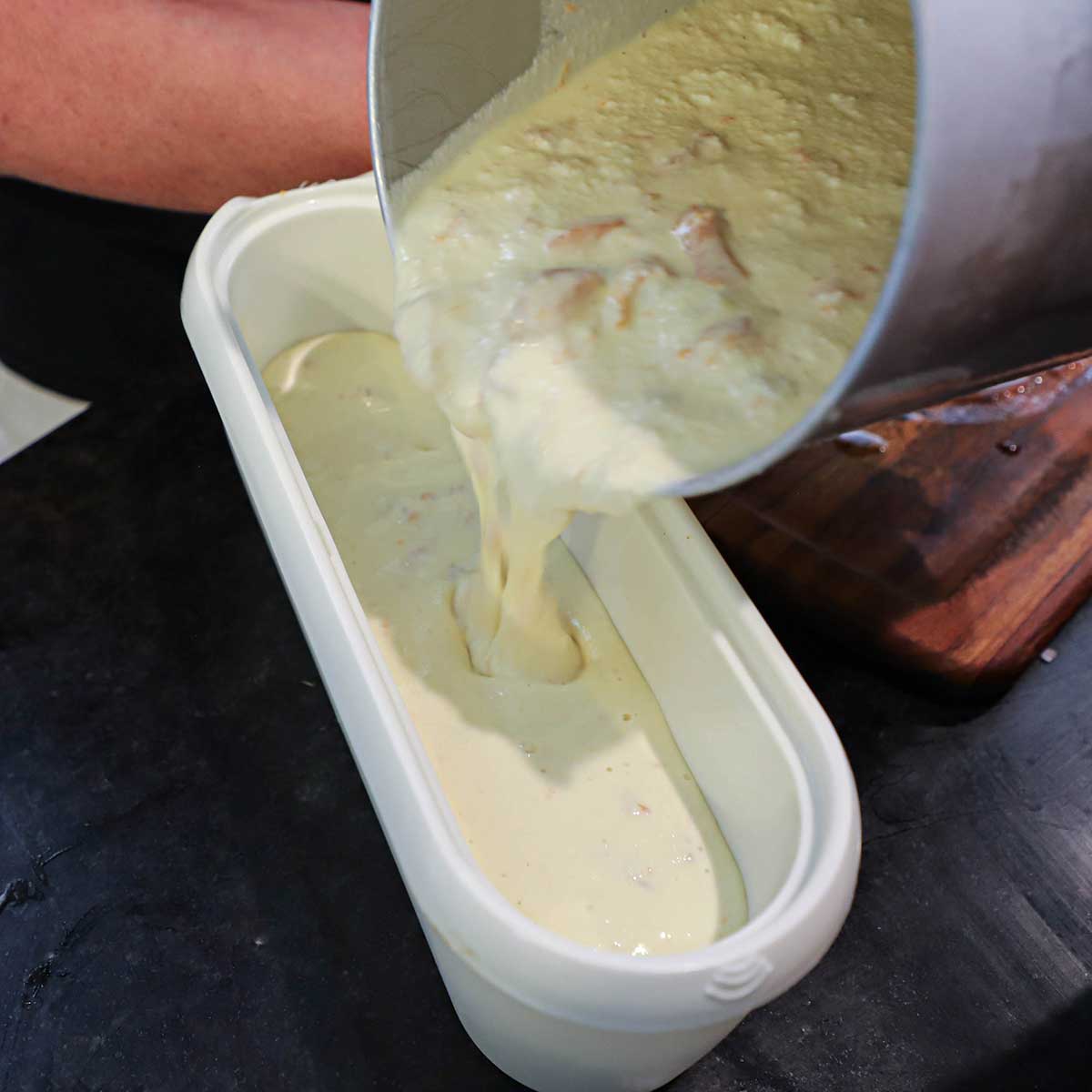 A person pouring soft, churned homemade peach ice cream from an ice cream canister into an oval ice cream container.