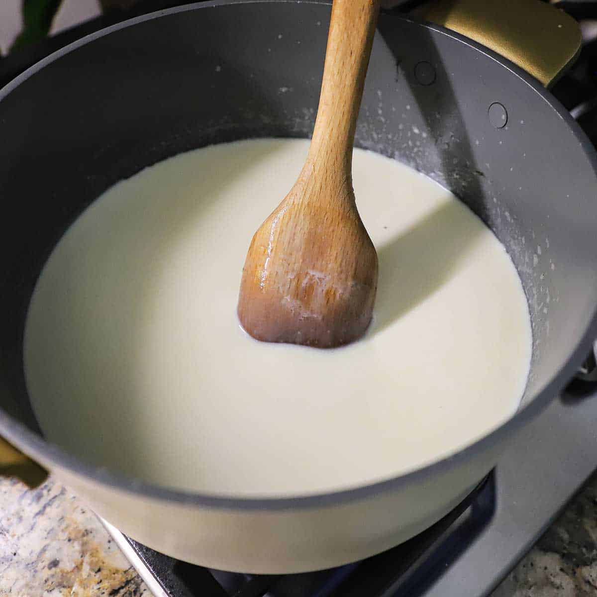 A person using a wooden spoon to stir cream and sugar in a large pot on a gas stove.