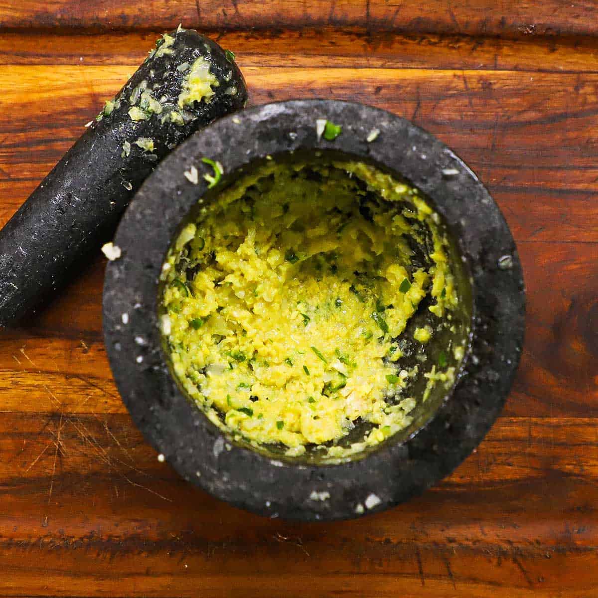 An overhead view of a mortar and pestle that contains freshly prepared ginger-garlic-green chili paste on a wooden cutting board.