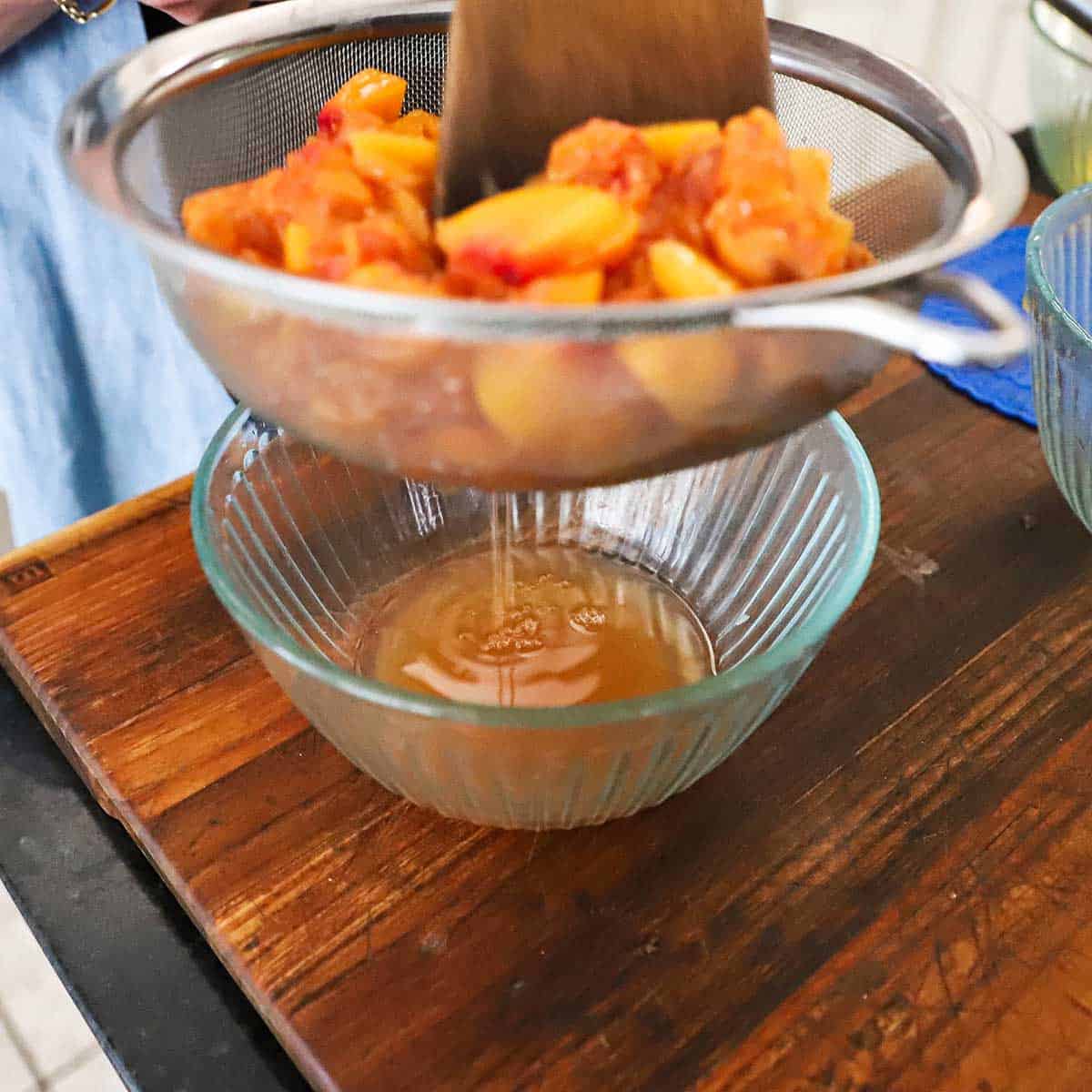 A person using a spatula to press on macerated peaches in a sieve with the juices running into a glass bowl beneath.