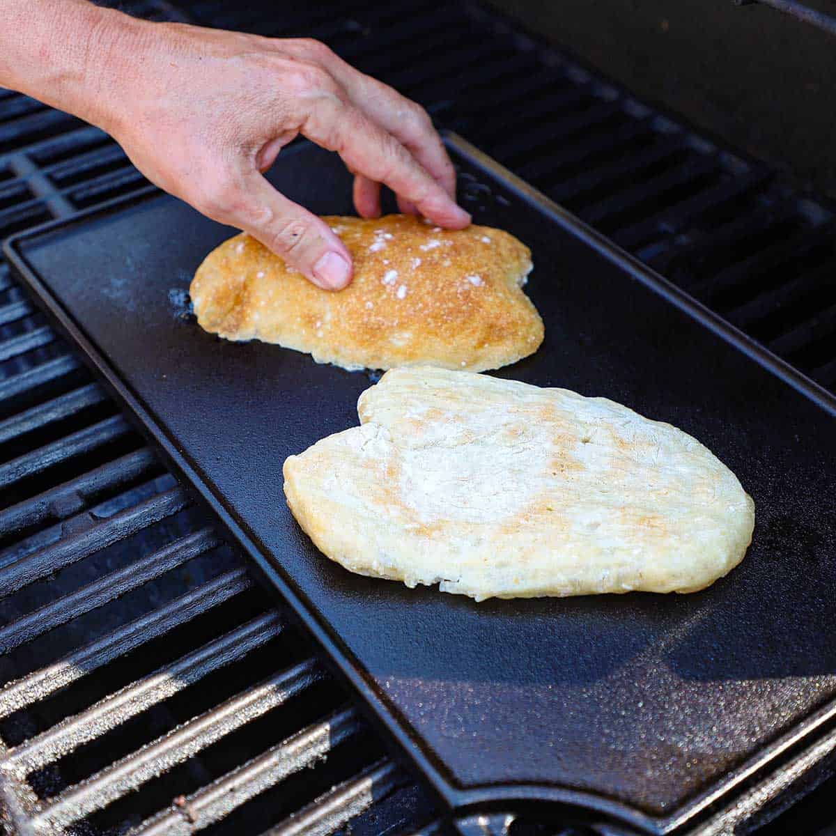 A person placing two halved ciabatta rolls cut side down onto a grill griddle on a gas grill.