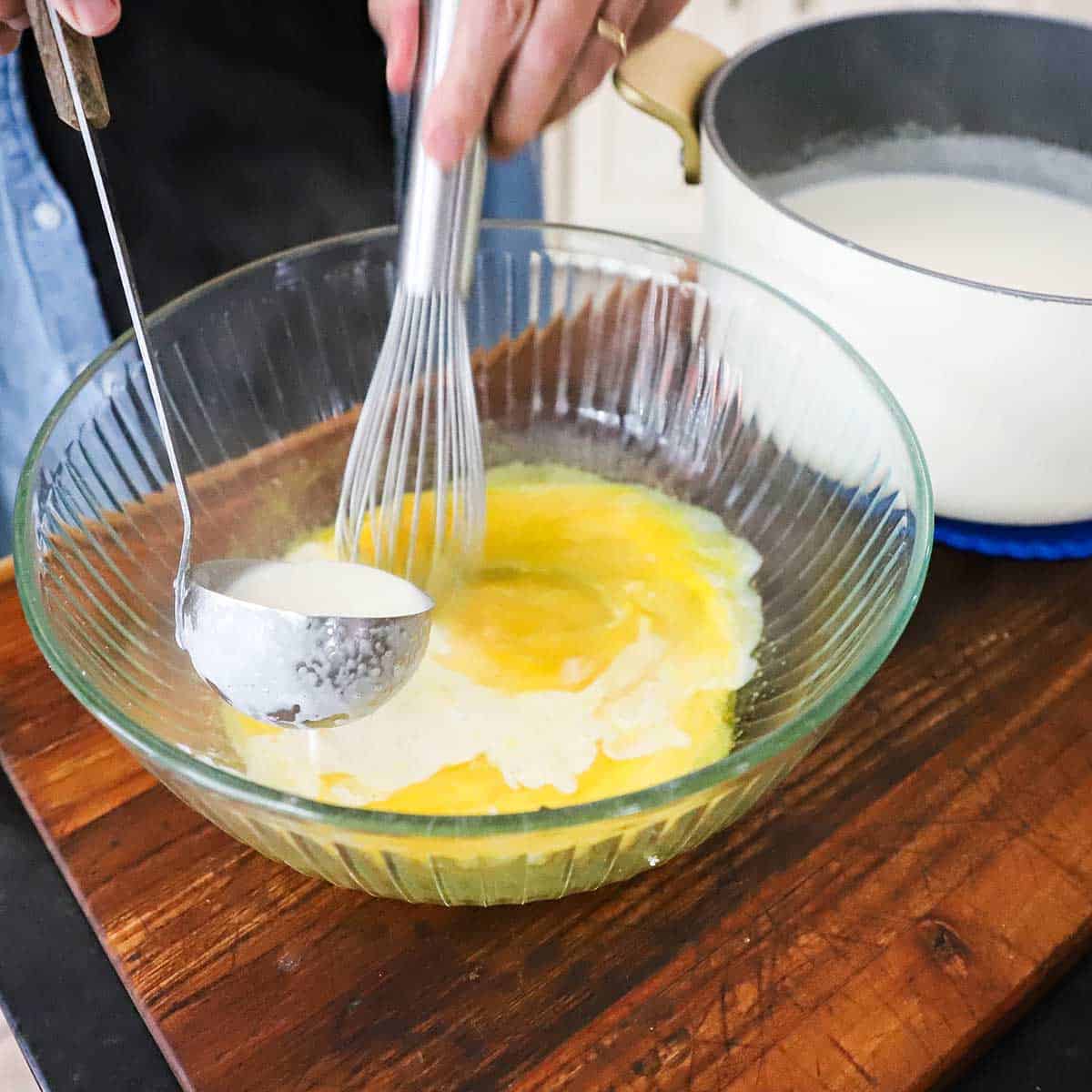A person using a whisk to vigorously stir warm cream into egg yolks in a large glass bowl on a wooden cutting board.