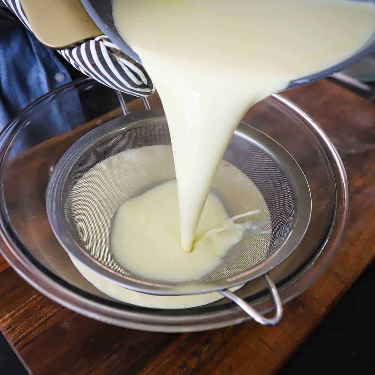 A person pouring vanilla custard from a large pot through a fine-mesh sieve into a large glass bowl on a wooden cutting board.