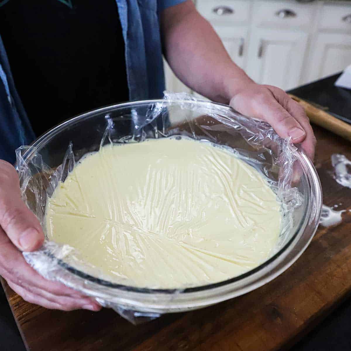 A person holding the sides of a large glass bowl that is filled with warm peach and vanilla custard with plastic wrap resting on the surface of the custard in the bowl.