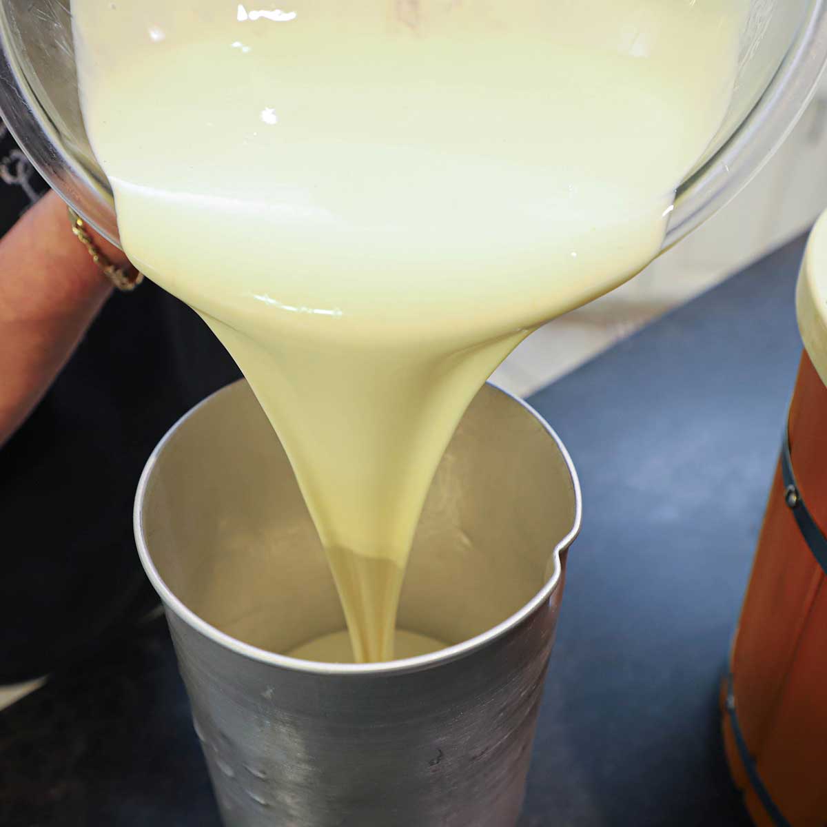 A person pouring chilled homemade peach ice cream custard from a large glass bowl into a metal ice cream canister on a kitchen counter.